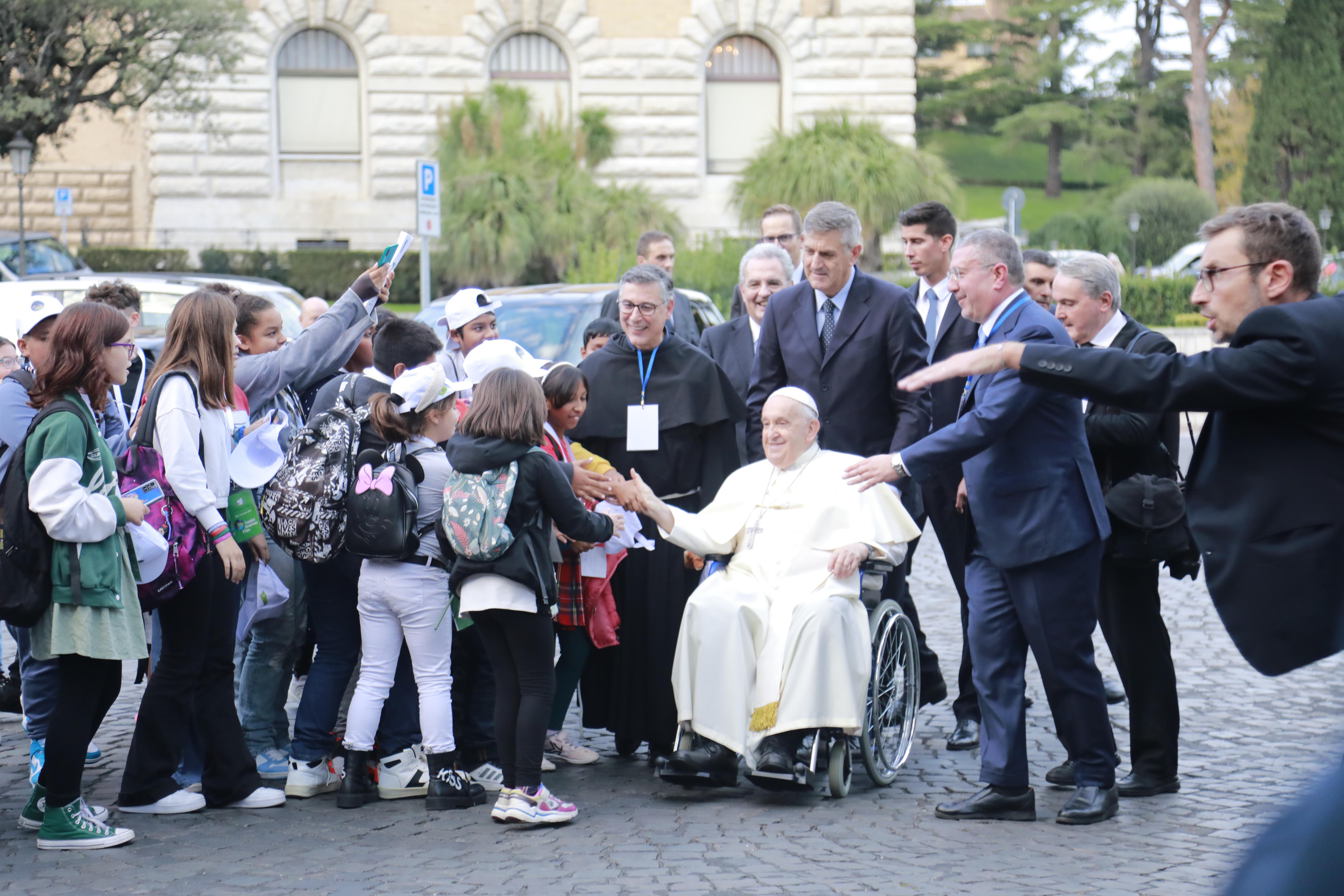 Paus Franciscus groet een groepje kinderen vanuit zijn rolstoel.
