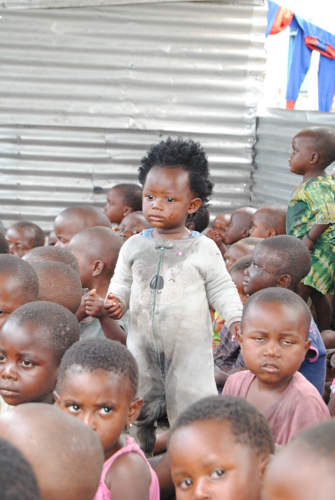 Kleutertje in een kruippak dat ooit wit was, te midden van een massa andere kinderen in het vluchtelingenkamp van zuster Marie-Leontine in Muganga.